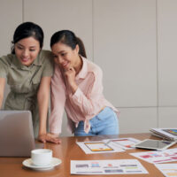 two-young-asian-female-coworkers-standing-desk-using-laptop