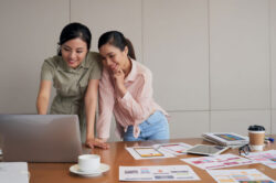 two-young-asian-female-coworkers-standing-desk-using-laptop